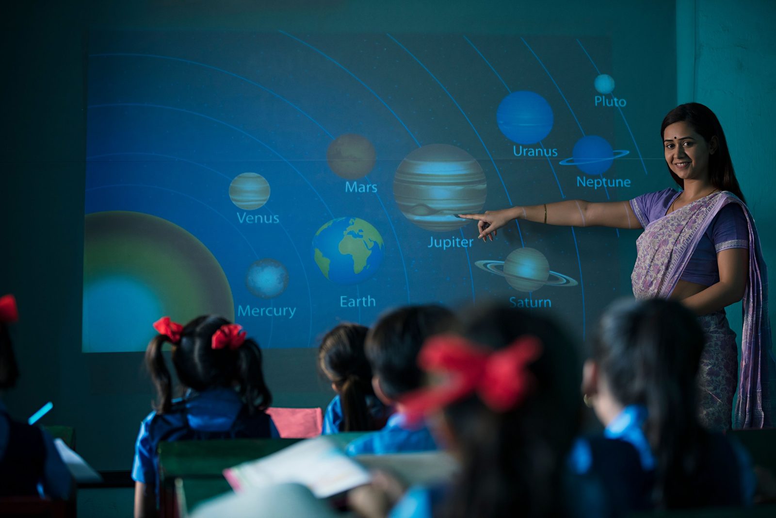 A teacher pointing to the solar system on a classroom smart screen while students follow the lesson